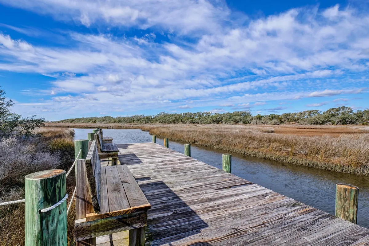 A wooden dock extends out over tranquil waters, surrounded by lush marshland and under a clear blue sky. The dock is the focal point, inviting activities such as fishing, kayaking, or simply enjoying peaceful moments by the water.
