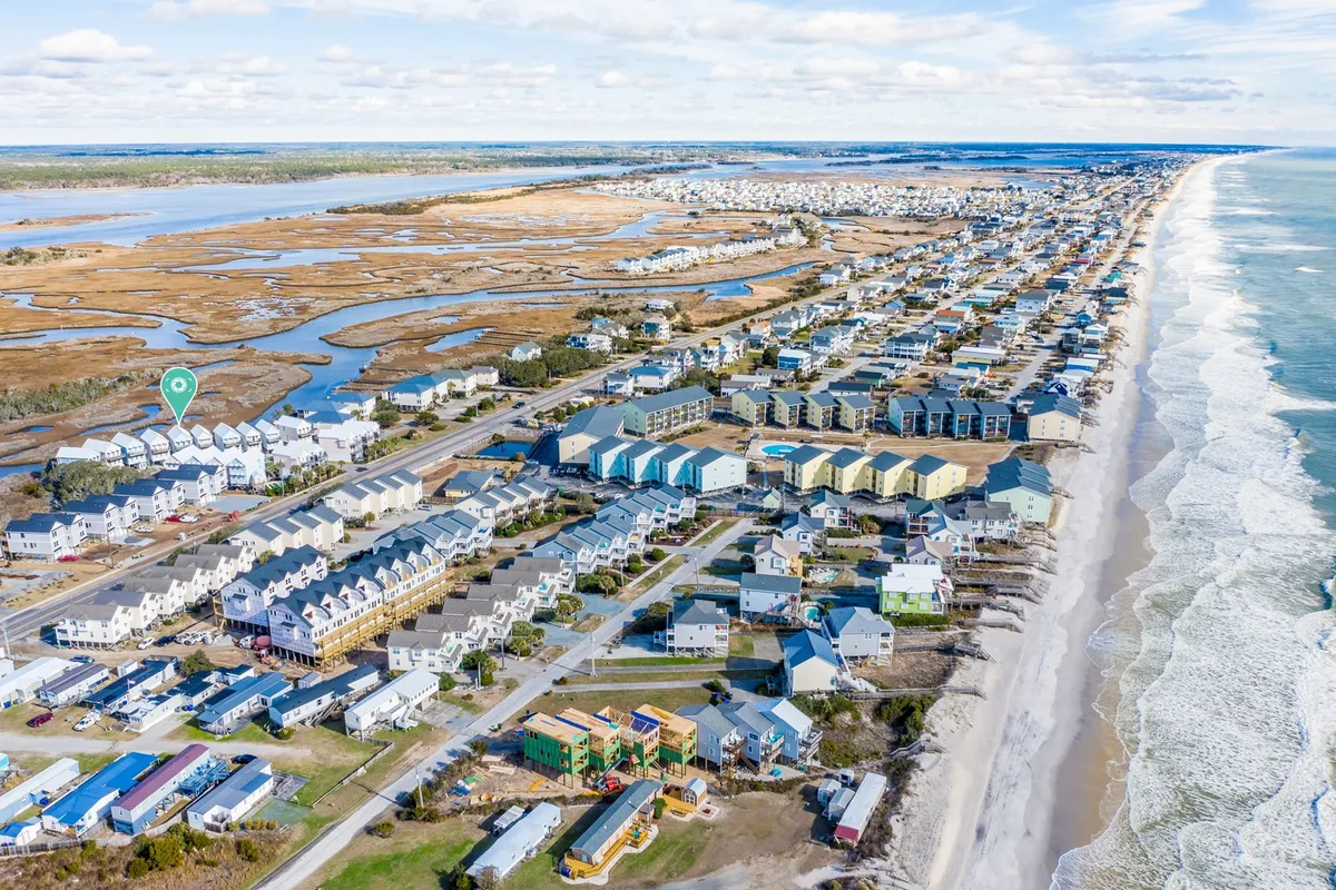 A broad exterior perspective featuring a beachfront with gentle ocean waves, marshland, and residential neighborhoods. The landscape shows the open, accessible coastline and a sense of community activity.