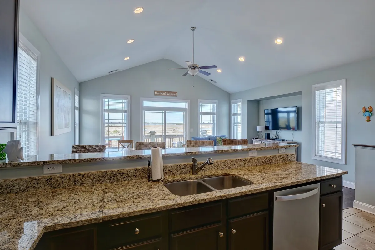 A spacious kitchen featuring a granite countertop, kitchen sink, large windows, ceiling fan, and a television. The space is designed for both functionality and gathering, with plenty of natural light and areas for family or group activities.