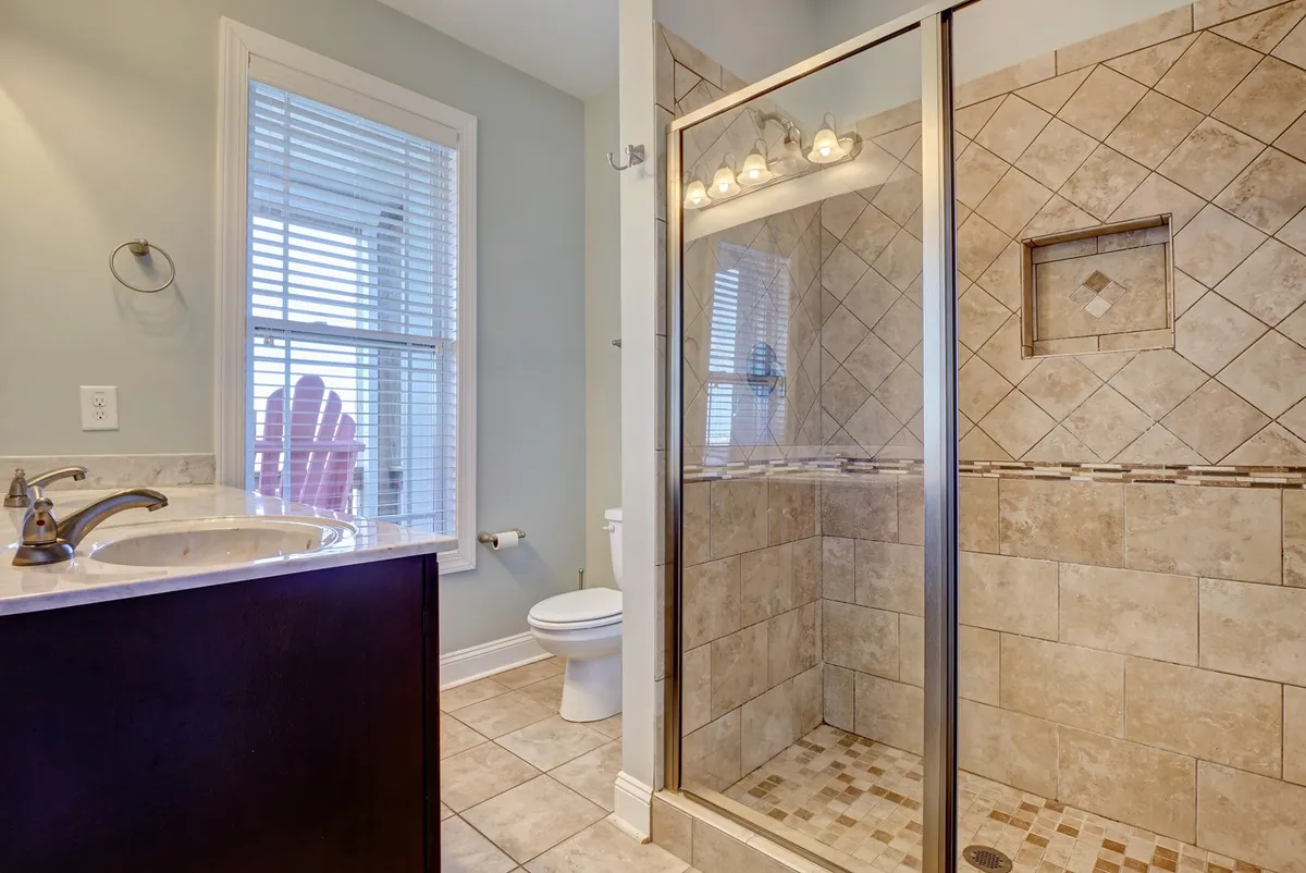 A modern bathroom with a sleek glass-door shower, a vanity with a sink, a window with blinds, and a toilet. The space is bright, clean, and inviting, offering spa-like amenities.