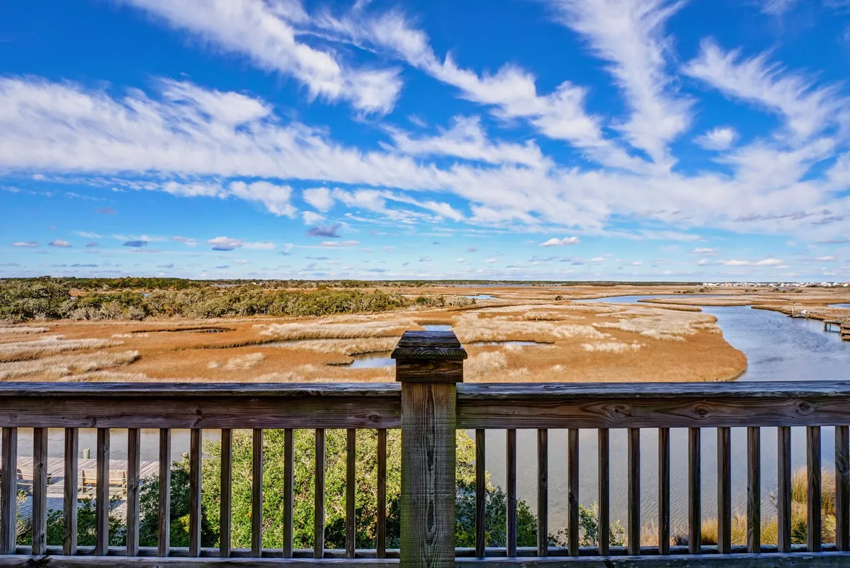 The image showcases a tranquil expanse of marshland with a winding river, framed by a dramatic sky, inviting guests to relax on the deck and take in the scenic natural beauty. Birdlife and the ever-changing light make the view ideal for sunset picnics, morning coffee, and peaceful nature observation.