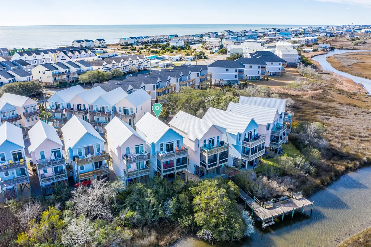 A scenic exterior view showcasing a picturesque row of beach houses facing the sea, a wooden dock stretching over the water, and vibrant greenery surrounding the homes. The image captures both the beauty of the coast and the inviting charm of the area.