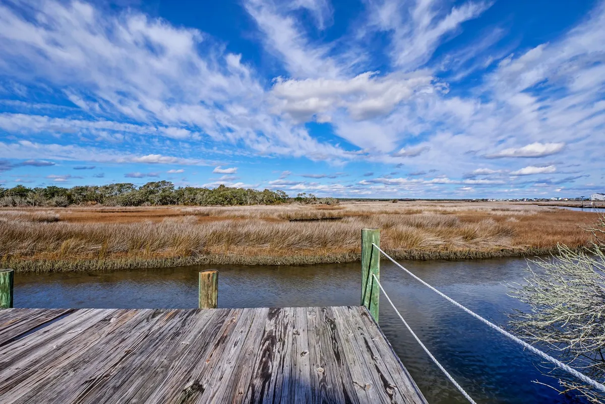 A tranquil view featuring a wooden dock extending over marshland, set against a sky dotted with clouds. This peaceful waterside scene suggests leisurely activities and year-round access to the area’s scenic waterways.
