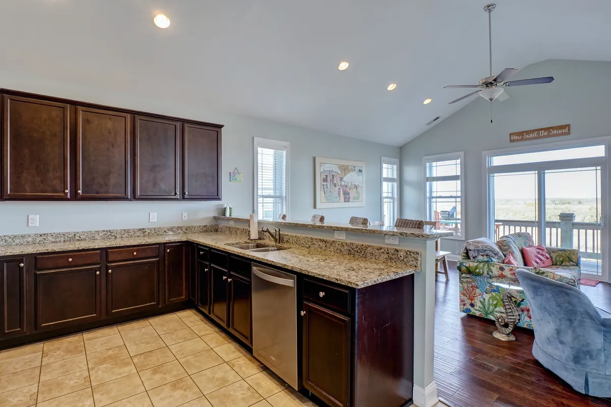 A spacious open-concept living, dining, and kitchen area featuring a granite countertop, dark wooden cabinets, a colorful sofa, and large windows. The layout encourages socializing, cooking, and enjoying the natural light and views beyond.